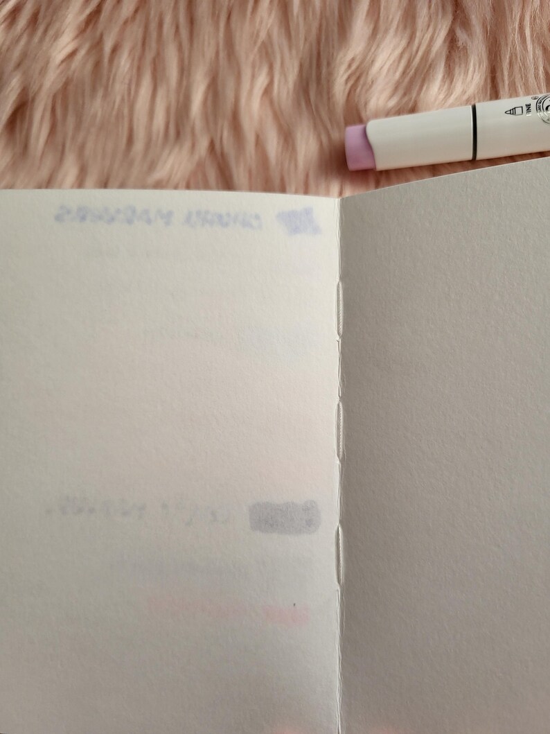 May include: Open blank notebook with stitched binding, pages are white. A white marker with a lavender cap rests on the top right corner. The background is a soft, pink, fuzzy material. Faint text is visible on the notebook pages.