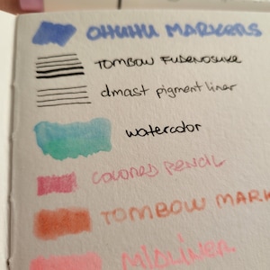 May include: A close-up of a white notepad with color swatches from various art supplies. The text "OHUHU MARKERS", "TOMBOW FUDENOSUKE", "dmast pigment liner", "watercolor", "colored pencil", "TOMBOW MARK", "Mildliner", and "copic MARK" are visible.