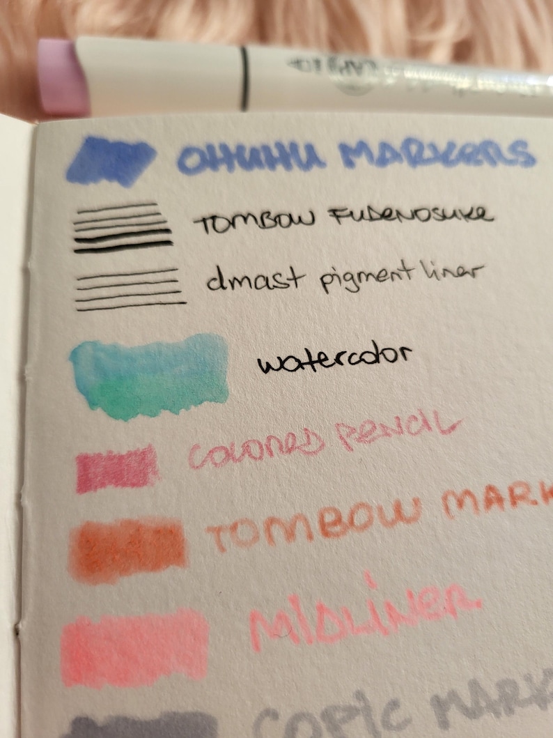May include: A close-up of a white notepad with color swatches from various art supplies. The text "OHUHU MARKERS", "TOMBOW FUDENOSUKE", "dmast pigment liner", "watercolor", "colored pencil", "TOMBOW MARK", "Mildliner", and "copic MARK" are visible.