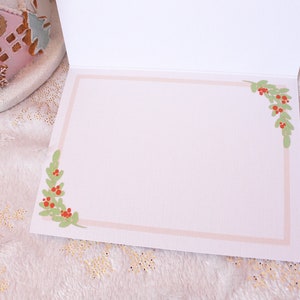 May include: Blank white card with a pink border and green holly sprigs with red berries in each corner.
