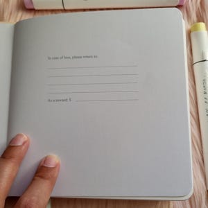 May include: Open notebook with lined pages, text reading "In case of loss, please return to:" and "As a reward: $".  White and pastel-colored markers surround the notebook on a pink, fuzzy surface.