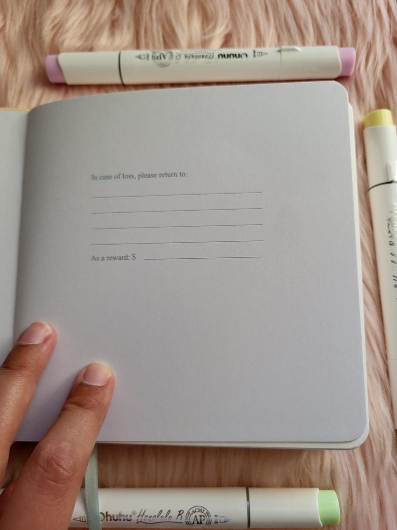 May include: Open notebook with lined pages, text reading "In case of loss, please return to:" and "As a reward: $".  White and pastel-colored markers surround the notebook on a pink, fuzzy surface.