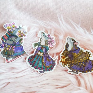 May include: Three mouse-shaped stickers with intricate details. Each mouse is wearing a different outfit, one with a floral crown, one with a purple dress, and one with a blue and gold dress.