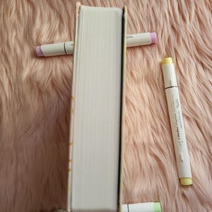 May include: A hardcover book propped up by four white markers with pastel-colored tips. The book's pages are white, and the spine is visible. The markers have pink, yellow, and green tips. The background is a soft, pink, fluffy surface.
