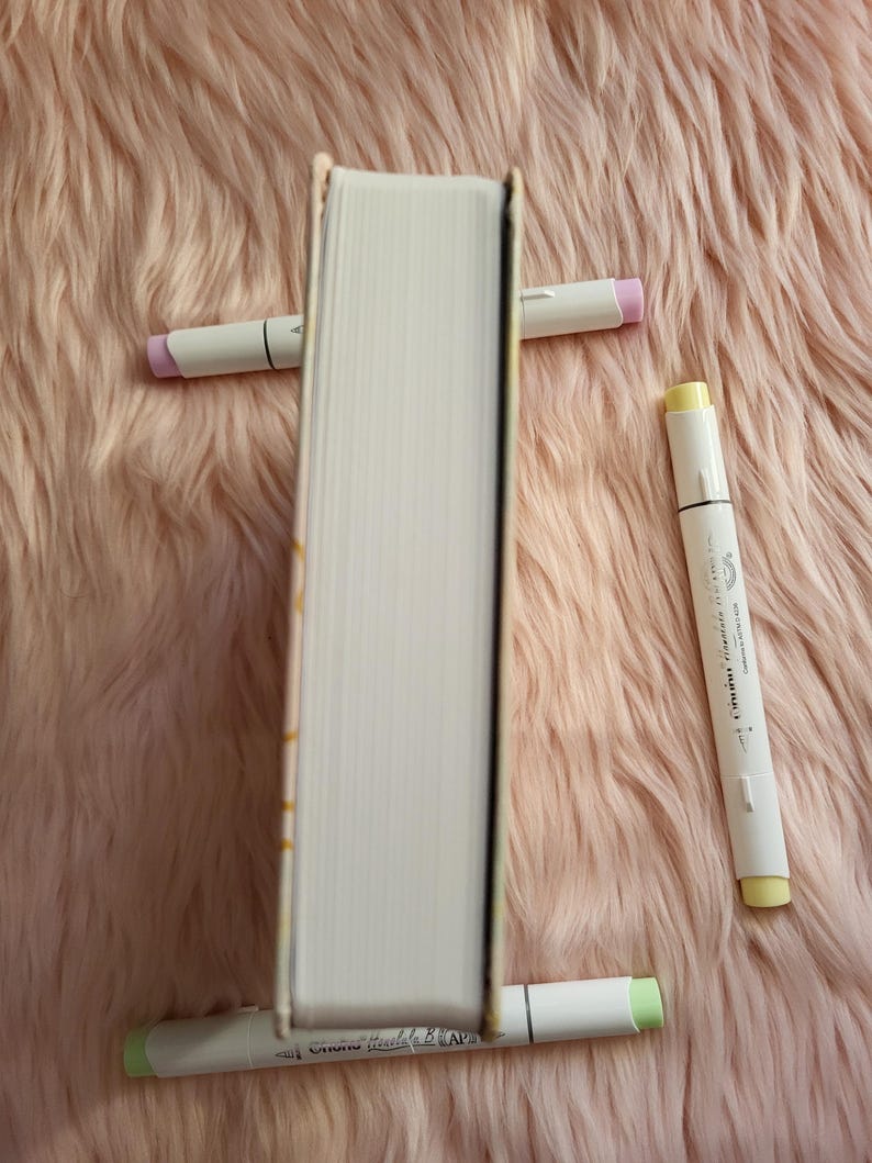 May include: A hardcover book propped up by four white markers with pastel-colored tips. The book's pages are white, and the spine is visible. The markers have pink, yellow, and green tips. The background is a soft, pink, fluffy surface.