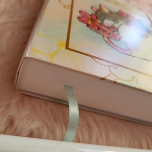 May include: A hardcover journal with a pastel floral illustration and a light blue ribbon bookmark. A white Ohuhu brush pen is in the foreground. The journal cover features a gold border and a whimsical design.