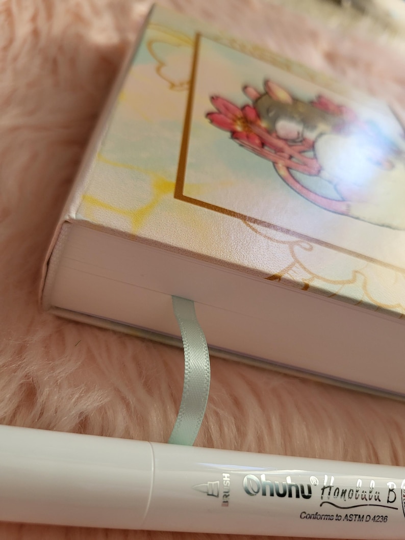 May include: A hardcover journal with a pastel floral illustration and a light blue ribbon bookmark. A white Ohuhu brush pen is in the foreground. The journal cover features a gold border and a whimsical design.