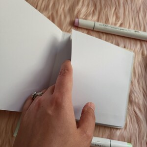 May include: An open, blank sketchbook with a hand holding the pages. Two art markers, one with a pink tip and the other with a green tip, are placed near the sketchbook. The background is a soft, pink, fuzzy material.