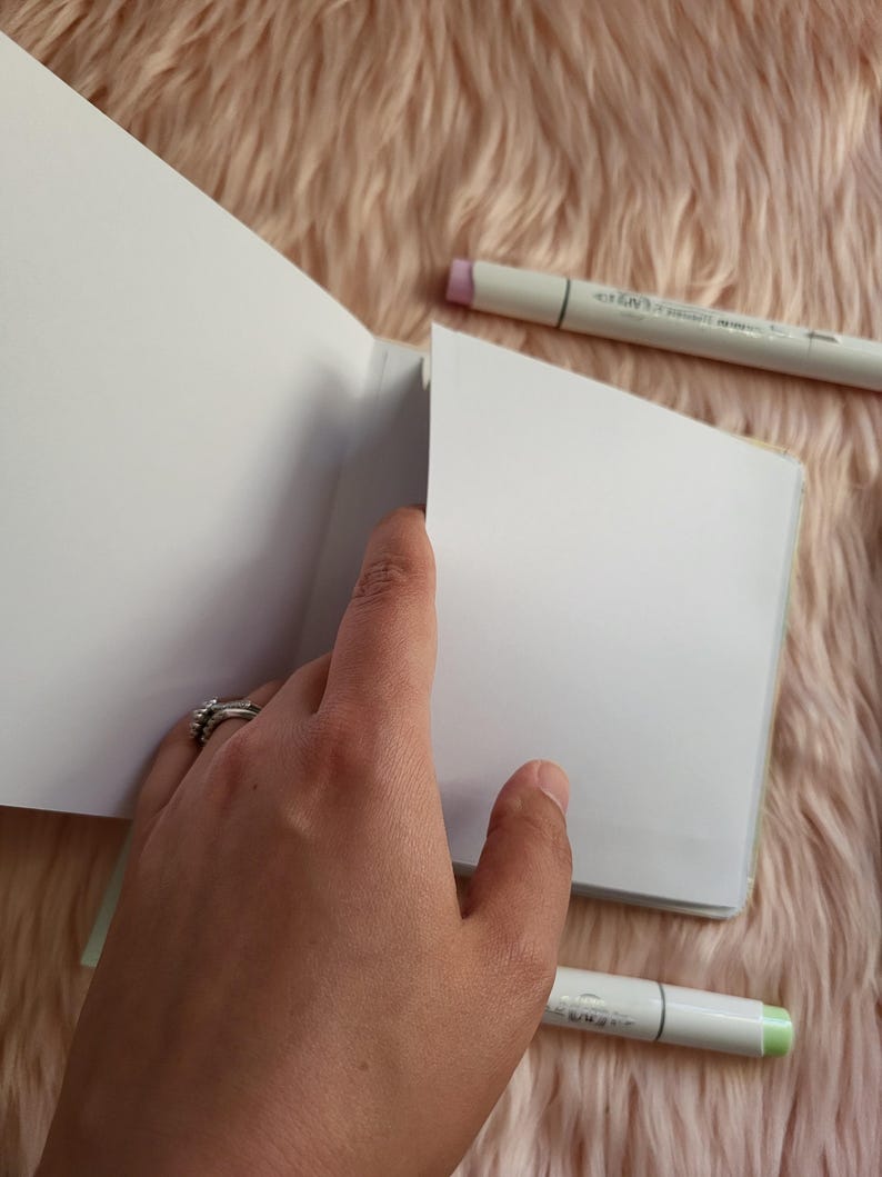 May include: An open, blank sketchbook with a hand holding the pages. Two art markers, one with a pink tip and the other with a green tip, are placed near the sketchbook. The background is a soft, pink, fuzzy material.