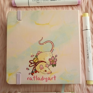 May include: A square art journal with a rat illustration and the text "ratladyart" is surrounded by three white art markers. The journal has a light pink cover with a watercolor design and pink ribbon closures.