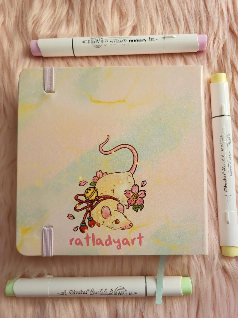 May include: A square art journal with a rat illustration and the text "ratladyart" is surrounded by three white art markers. The journal has a light pink cover with a watercolor design and pink ribbon closures.
