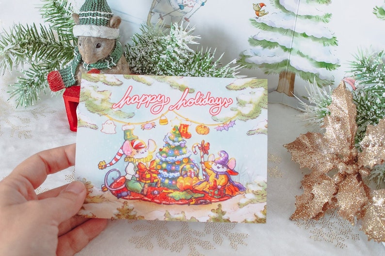 May include: A Christmas card with a blue background featuring three mice on a red sled. The mice are pulling a decorated Christmas tree. The card says "Happy Holidays" in pink lettering.