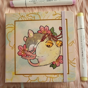 May include: A square journal with a floral design and a sleeping rat illustration, surrounded by pink flowers and gold bells. Three white Ohuhu Honolulu B markers with pink, yellow, and green tips are arranged around the journal.