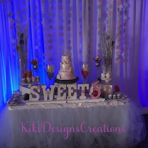 May include: A white table with a white tulle tablecloth and a dessert table decorated with a pink and white cake, white wooden letters spelling "SWEET 16", and various other decorations. The backdrop is a white curtain with blue lighting and snowflake decorations.
