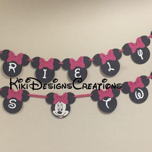 May include: A black and pink Minnie Mouse themed banner with the letters "RILEY IS" in white on black circles. Each circle has a pink bow on top.