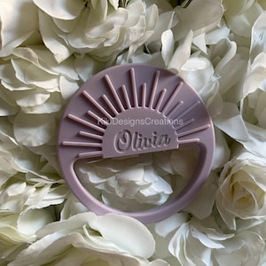 May include: A light purple, circular bottle opener with a sunburst design and the name "Olivia" engraved. The text "KikiDesignsCreations" is also visible. The opener has a handle and is set against a backdrop of white flowers.