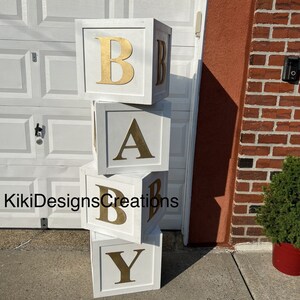 Puede incluir: Bloques de madera blancos apilados para deletrear "BABY" con letras doradas. Los bloques est&aacute;n sobre una superficie de concreto gris frente a una puerta de garaje blanca.