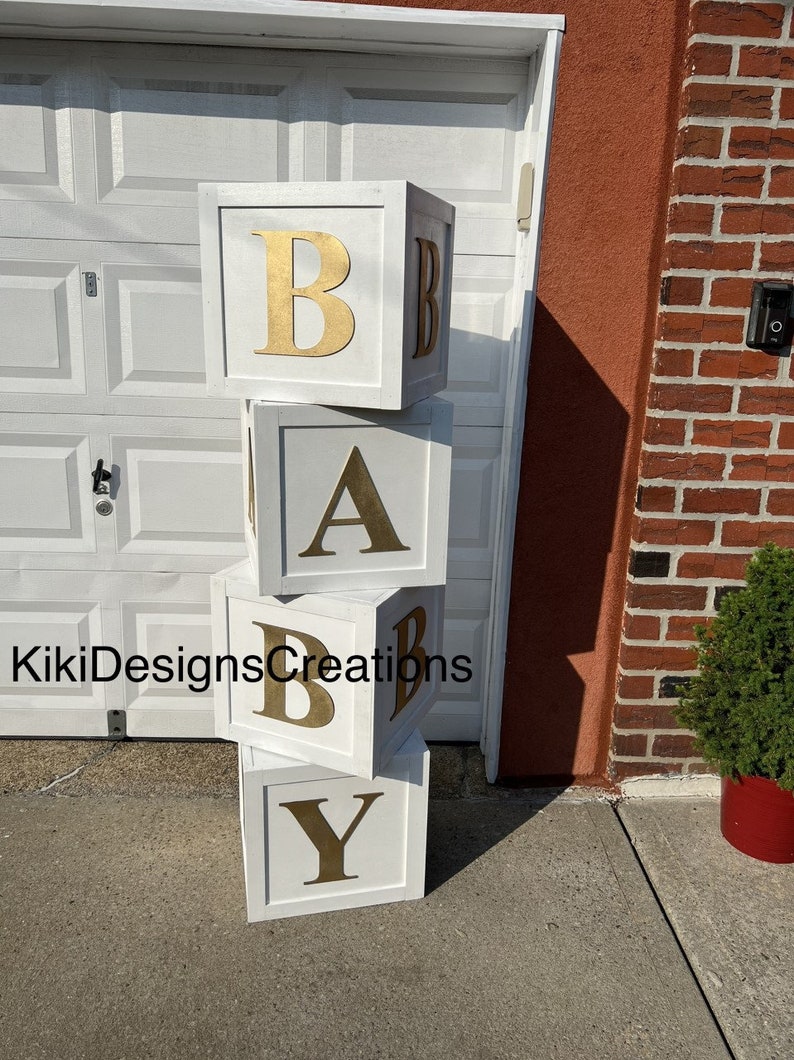 Puede incluir: Bloques de madera blancos apilados para deletrear "BABY" con letras doradas. Los bloques est&aacute;n sobre una superficie de concreto gris frente a una puerta de garaje blanca.