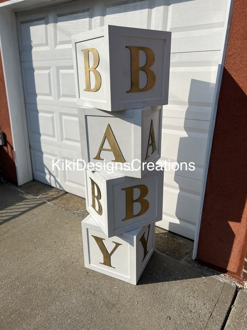 Puede incluir: Bloques de madera blancos apilados para deletrear "BABY" con letras doradas. Los bloques est&aacute;n pintados de blanco y tienen un aspecto ligeramente desgastado.