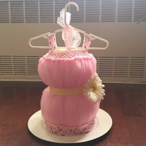 Puede incluir: Un pastel de pa&ntilde;ales para baby shower de tul rosa decorado con una cinta amarilla, una flor blanca y una cinta con estampado floral. El pastel est&aacute; en un plato blanco y est&aacute; colgado de una percha blanca.