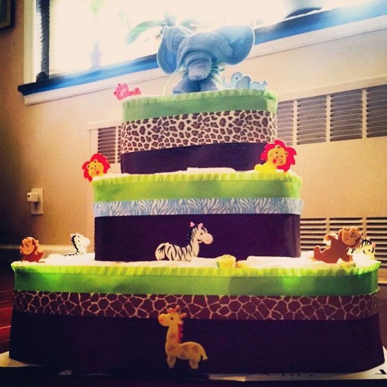 Puede incluir: Un pastel de pa&ntilde;ales de varios niveles decorado con figuritas de elefante azul, cebra y jirafa. El pastel est&aacute; envuelto en papel y cintas con patrones marrones, verdes y azules.