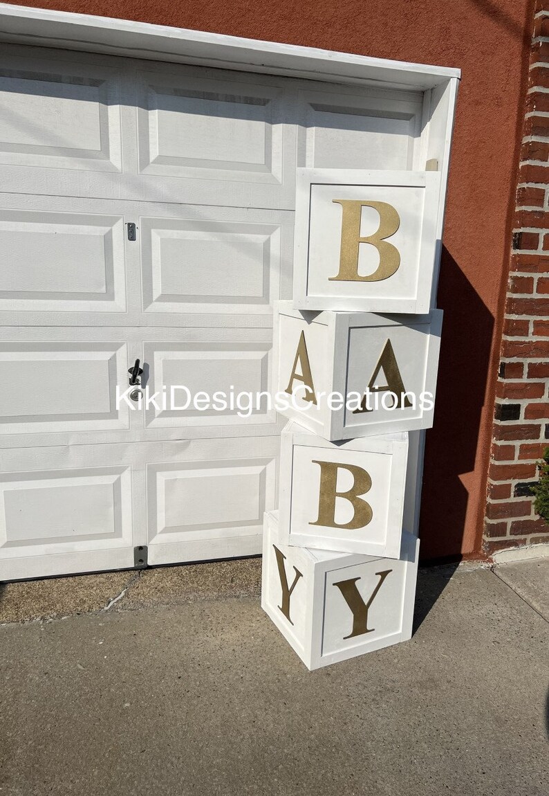 Puede incluir: Una pila de cuatro bloques de madera blancos con letras doradas que deletrean la palabra "BABY".