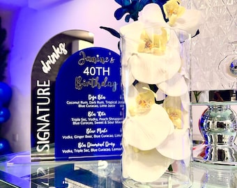 May include: A clear glass vase filled with white orchid petals and blue flowers. A sign with the words "Signature Drinks" and "Janine's 40th Birthday" is in the background, listing cocktail names and ingredients.