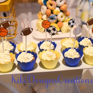 May include: A display of cupcakes with white frosting, each topped with a sports-themed cutout: a basketball, baseball, soccer ball, and football. The cupcakes are in blue and yellow cupcake liners, arranged on a white platter.
