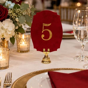 May include: A deep red, scalloped-edge table number card featuring the gold-colored numeral 5 in the center. The card is held upright by a small, polished gold-toned metal stand with a circular base, positioned on a white tablecloth.