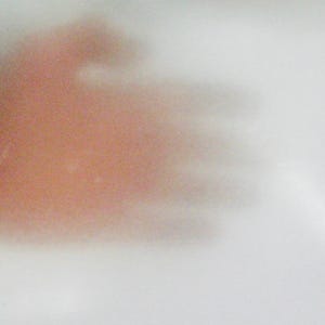 May include: A translucent white material with a soft, blurred, and indistinct reddish-orange shape visible behind the surface. The texture appears smooth and thin, creating a diffused effect that obscures the details of the object behind it.