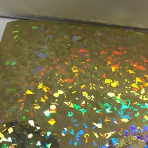 K&ouml;nnte beinhalten: Ein Bogen aus schillernder Goldfolie mit einem Streumuster aus bunten, geometrischen Formen. Die Formen reflektieren ein Farbspektrum, darunter Blau, Gr&uuml;n, Gelb und Orange. Die Folie hat eine gl&auml;nzende, reflektierende Oberfl&auml;che.