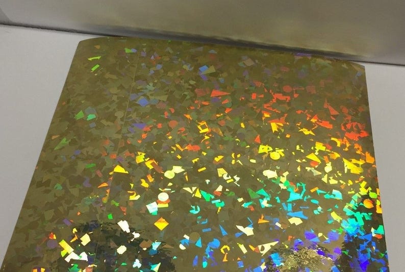 K&ouml;nnte beinhalten: Ein Bogen aus schillernder Goldfolie mit einem Streumuster aus bunten, geometrischen Formen. Die Formen reflektieren ein Farbspektrum, darunter Blau, Gr&uuml;n, Gelb und Orange. Die Folie hat eine gl&auml;nzende, reflektierende Oberfl&auml;che.