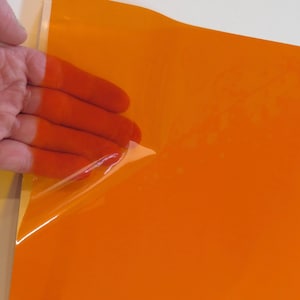 May include: A rectangular sheet of vibrant orange adhesive vinyl material with a smooth, glossy finish. The top corner of the orange film is partially peeled back, revealing a clear, transparent backing layer underneath the colored surface.