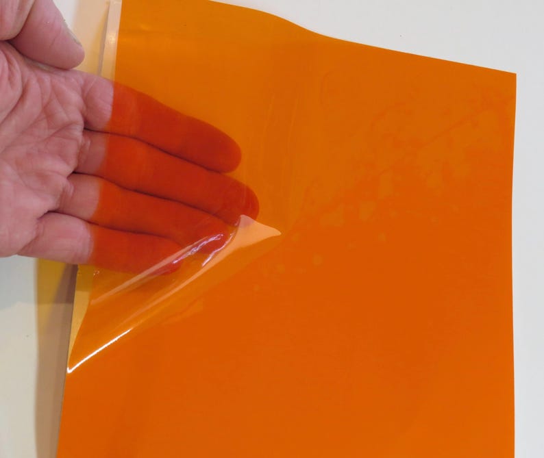 May include: A rectangular sheet of vibrant orange adhesive vinyl material with a smooth, glossy finish. The top corner of the orange film is partially peeled back, revealing a clear, transparent backing layer underneath the colored surface.
