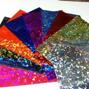 K&ouml;nnte beinhalten: F&auml;cherf&ouml;rmig angeordnete, farbenfrohe Hologrammfolien. Die Folien zeigen verschiedene Farben, darunter Rot, Blau und Silber. Jede Folie hat eine gl&auml;nzende, reflektierende Oberfl&auml;che mit einem konfettiartigen Muster. Anordnung in einem Halbkreis.