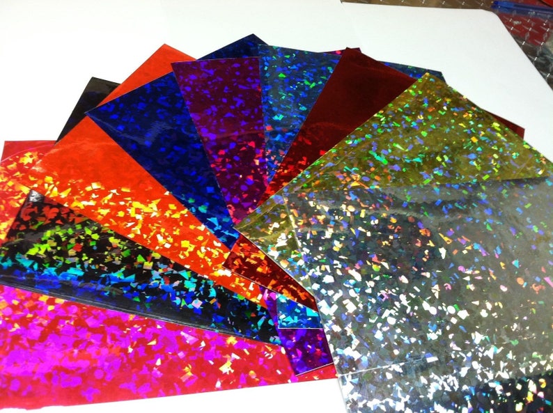 K&ouml;nnte beinhalten: F&auml;cherf&ouml;rmig angeordnete, farbenfrohe Hologrammfolien. Die Folien zeigen verschiedene Farben, darunter Rot, Blau und Silber. Jede Folie hat eine gl&auml;nzende, reflektierende Oberfl&auml;che mit einem konfettiartigen Muster. Anordnung in einem Halbkreis.