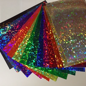 K&ouml;nnte beinhalten: Eine Sammlung von schillernden holografischen Bl&auml;ttern in verschiedenen Farben, darunter Schwarz, Lila, Rot, Orange, Gelb, Gr&uuml;n, Blau und Silber. Jedes Blatt hat eine reflektierende, strukturierte Oberfl&auml;che mit einem glitzerartigen Effekt, der einen Regenbogen von Farben erzeugt.