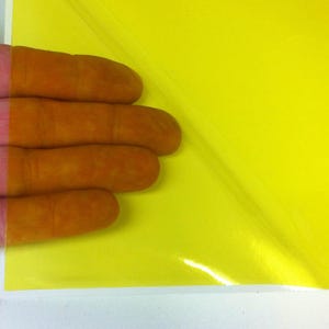 May include: A sheet of bright yellow adhesive vinyl film with a glossy finish. The material is partially peeled back from its white paper backing, revealing the vibrant yellow color and the smooth, reflective surface of the adhesive layer.