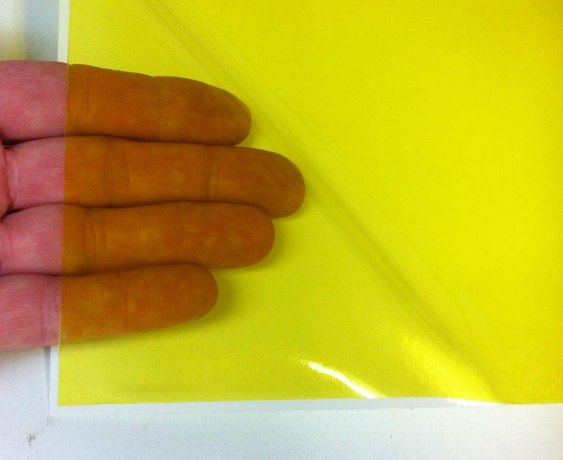 May include: A sheet of bright yellow adhesive vinyl film with a glossy finish. The material is partially peeled back from its white paper backing, revealing the vibrant yellow color and the smooth, reflective surface of the adhesive layer.