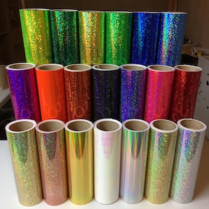 May include: A collection of twenty cylindrical rolls of adhesive vinyl arranged in three rows. The rolls feature various finishes including holographic, glitter, and solid metallic colors such as lime, green, blue, orange, red, pink, gold, and silver.