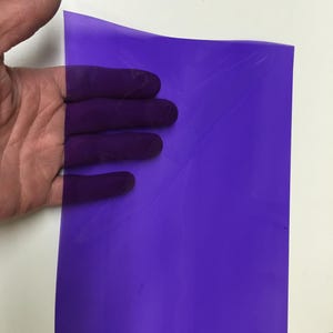 May include: A rectangular sheet of translucent purple plastic film. The material is thin, flexible, and smooth, allowing light to pass through while displaying a deep violet hue. The surface is flat with subtle creases visible across the sheet.