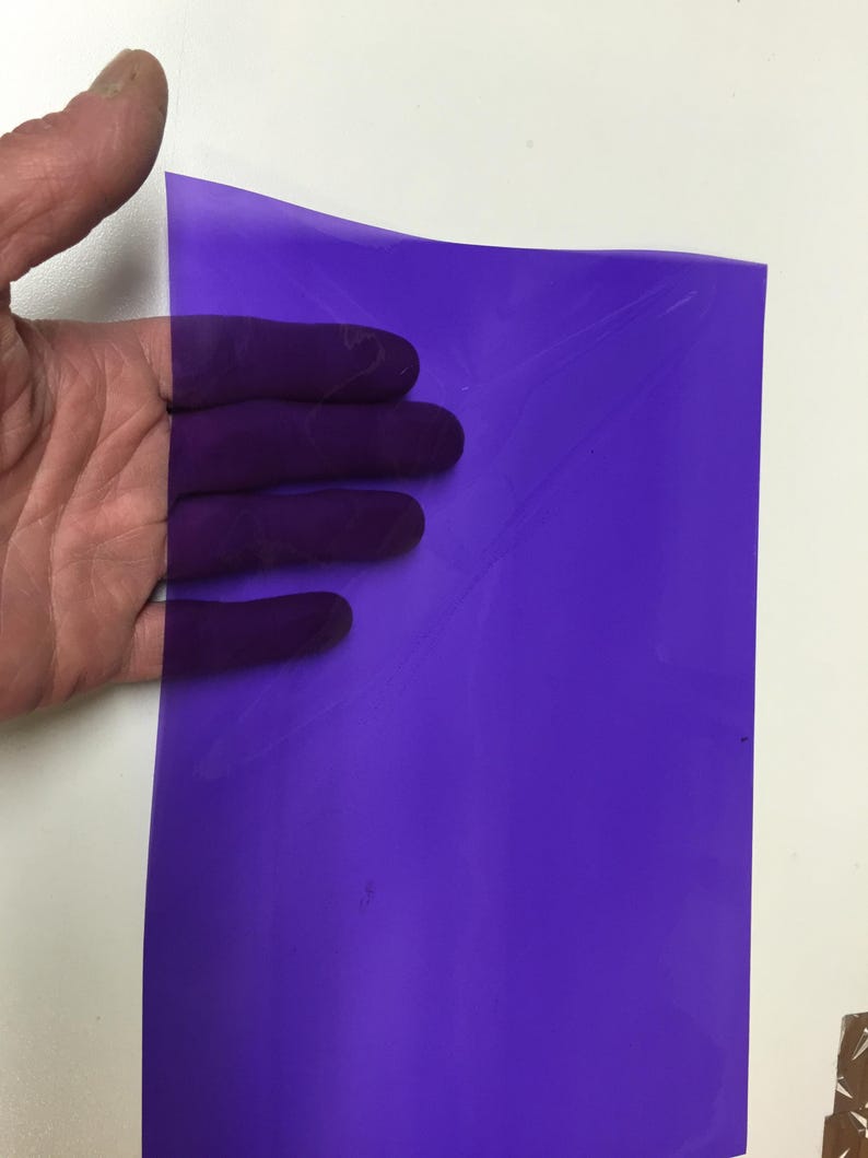 May include: A rectangular sheet of translucent purple plastic film. The material is thin, flexible, and smooth, allowing light to pass through while displaying a deep violet hue. The surface is flat with subtle creases visible across the sheet.