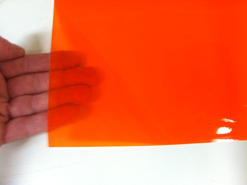 May include: A rectangular sheet of translucent orange plastic film with a smooth, glossy surface. The material is semi-transparent, allowing light to pass through while maintaining a vibrant, solid orange color across the entire flat surface.