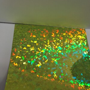 K&ouml;nnte beinhalten: Ein Bogen irisierendes, limettengr&uuml;nes holografisches Papier. Die Oberfl&auml;che ist mit kleinen, unregelm&auml;&szlig;igen Formen bedeckt, die mehrere Farben reflektieren, darunter Orange, Gelb und Blau. Das Papier ist teilweise vor einem wei&szlig;en Hintergrund sichtbar.