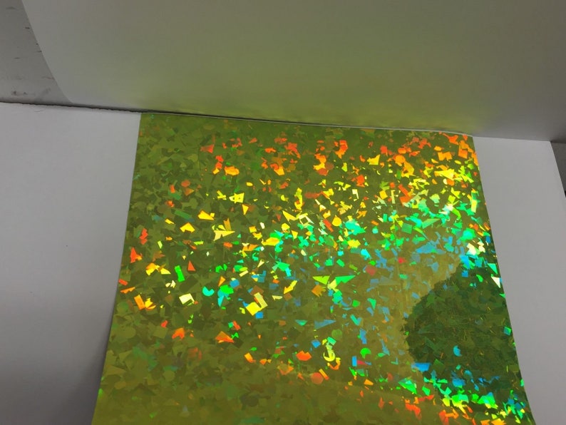 K&ouml;nnte beinhalten: Ein Bogen irisierendes, limettengr&uuml;nes holografisches Papier. Die Oberfl&auml;che ist mit kleinen, unregelm&auml;&szlig;igen Formen bedeckt, die mehrere Farben reflektieren, darunter Orange, Gelb und Blau. Das Papier ist teilweise vor einem wei&szlig;en Hintergrund sichtbar.
