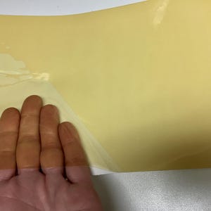 May include: A rectangular sheet of pale yellow adhesive material with a smooth, semi-glossy surface. The top left corner of the sheet is partially peeled back, revealing a thin, transparent backing layer underneath the opaque yellow film.