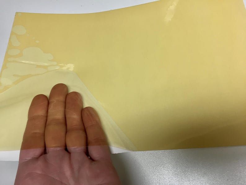 May include: A rectangular sheet of pale yellow adhesive material with a smooth, semi-glossy surface. The top left corner of the sheet is partially peeled back, revealing a thin, transparent backing layer underneath the opaque yellow film.