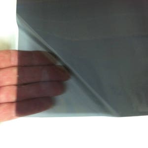 May include: A flexible, semi-transparent sheet of dark gray window tint film. The material is thin and smooth, allowing light to pass through while darkening the view. The film is shown partially peeled back to reveal its translucent quality.
