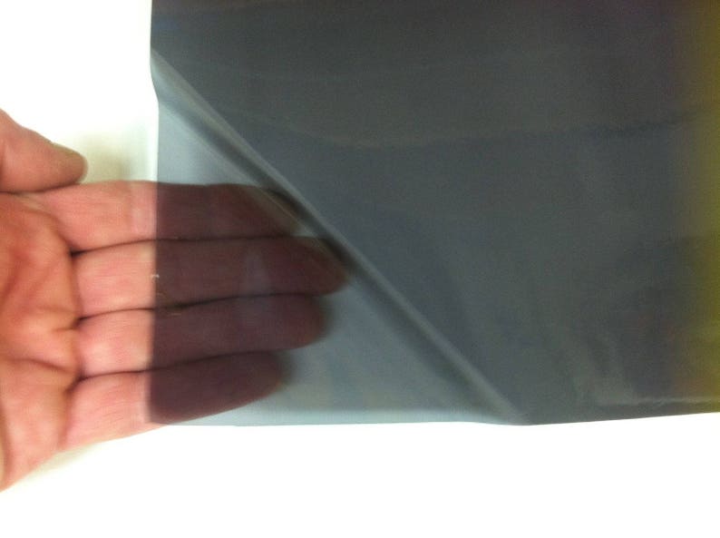 May include: A flexible, semi-transparent sheet of dark gray window tint film. The material is thin and smooth, allowing light to pass through while darkening the view. The film is shown partially peeled back to reveal its translucent quality.