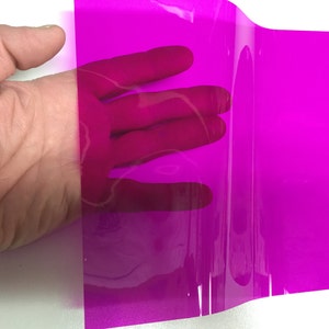 May include: A rectangular sheet of transparent magenta plastic film. The material is smooth, flexible, and semi-transparent, allowing light to pass through while casting a vibrant pink hue over the surface behind the sheet.